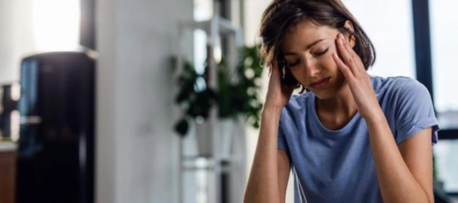 Mulher branca com dor de cabeça, sentada na cama com os olhos fechados e as mãos na cabeça