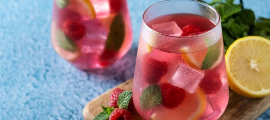 copo de vidro com líquido rosa, folhas de hortelã, frutas vermelhas e cubos de gelo e limão ao lado