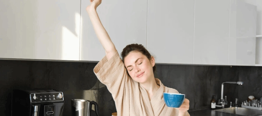 mulher feliz na cozinha usando roupão aproveitando os benefícios da cafeína com uma xícara na mão