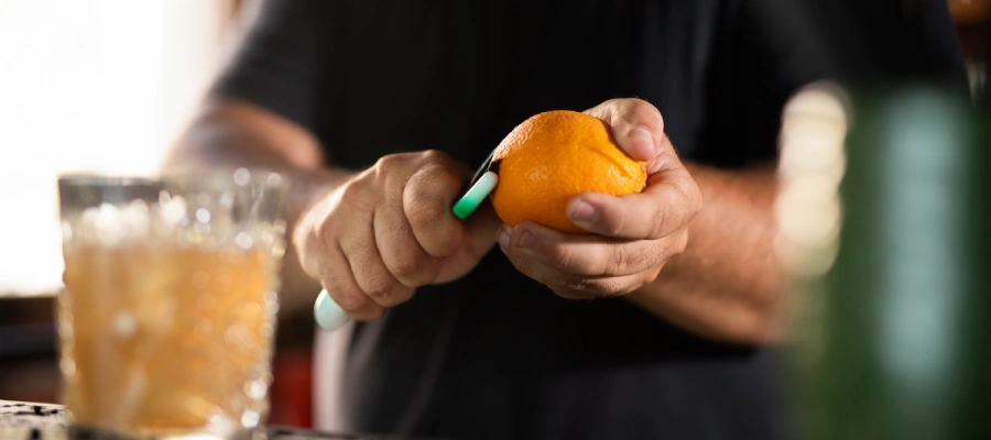 homem partindo uma laranja ao meio enquanto prepara um drink em um copo de vidro.