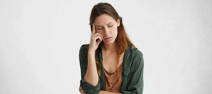 Mulher cansada em pé contra uma parede branca com os olhos fechados, segurando o dedo indicador na testa