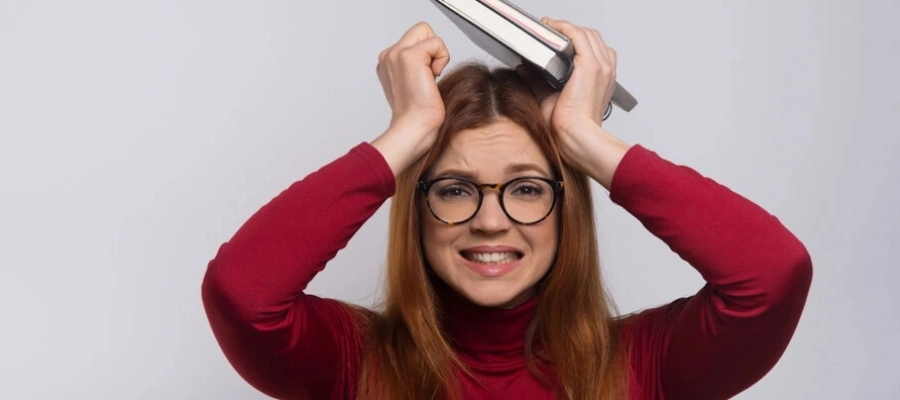 mulher com blusa vermelha e óculos segurando livro e caderno com as mãos na cabeça e expressão de dúvida